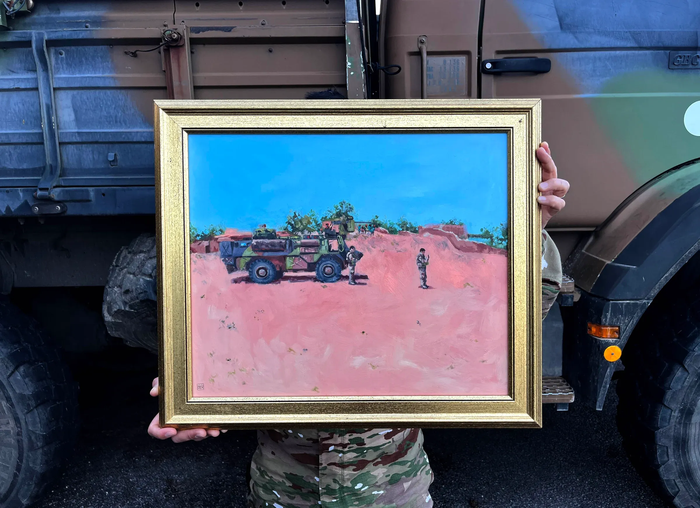 Petit tableau encadré, tenu à bout de bras devant un camion militaire : un véhicule isolé sur une piste de sable rouge