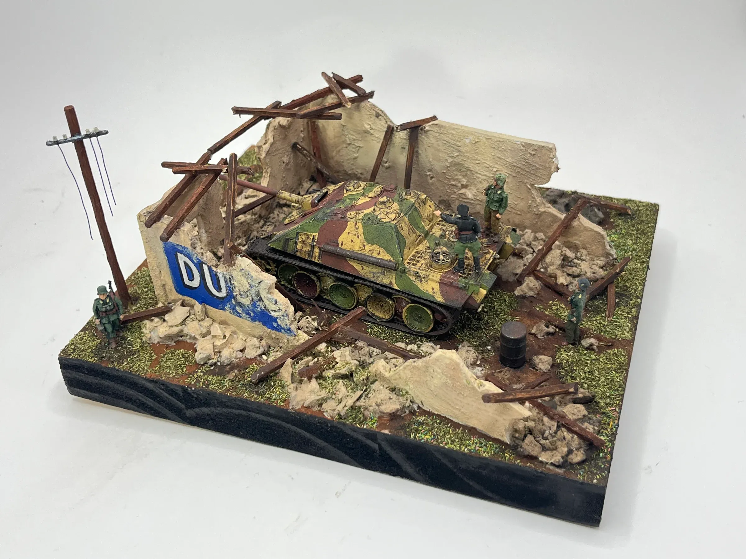 Diorama : un char camouflé dans les ruines d’un village, devant un mur peint d’une réclame