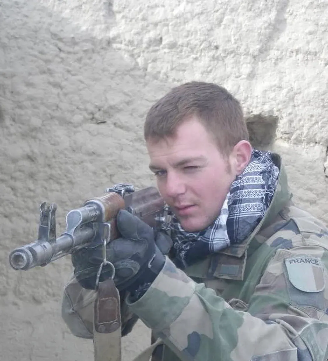 Photo de terrain : un jeune soldat en tenue camouflée, chèche autour du cou, épaule un fusil AK