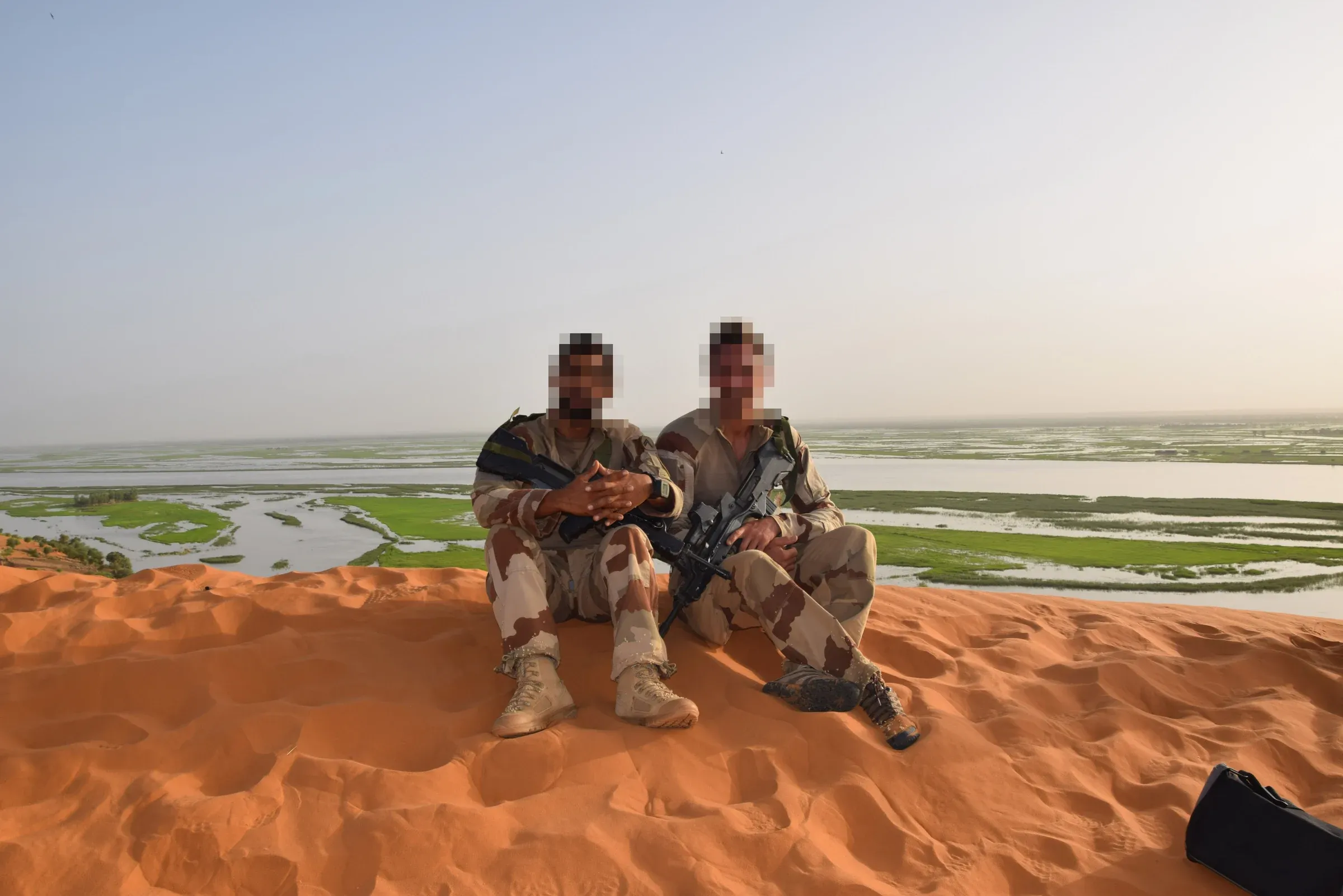 Photo de terrain : deux soldats assis sur une dune au-dessus d’un fleuve, visages floutés
